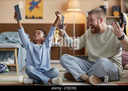 Il bambino nero entusiasta che si diverte a giocare vince giocando al telefono cellulare con il padre adottivo felice mentre è seduto sul pavimento nella stanza dei bambini Foto Stock