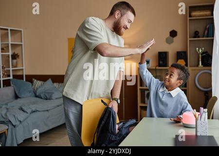 Vista laterale del allegro padre adottivo che dà cinque anni al bambino di colore che sostiene il figlio nel successo accademico, mentre aiuta il bambino a mettere in valigia la borsa della scuola in chi Foto Stock