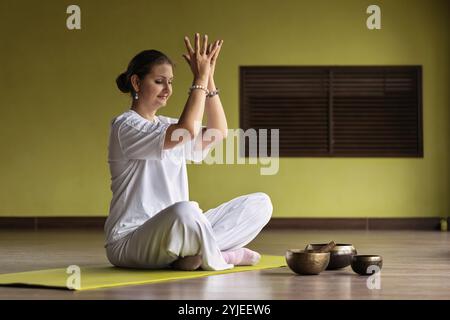 Donna che pratica kundalini yoga, meditando seduto in comoda posizione sul tappetino in camera e facendo mudra di loto con le mani in alto, vista a tutta lunghezza Foto Stock