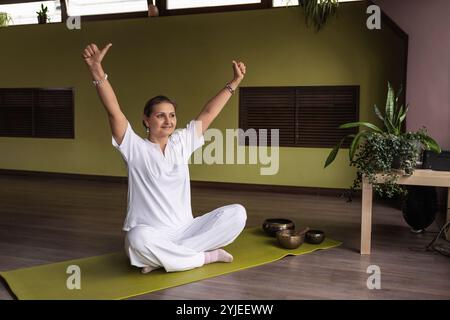Donna soddisfatta che pratica kundalini yoga, seduta sul tappetino in comoda posizione di loto con le braccia sollevate facendo ego o mudra di personalità, esercitandosi Foto Stock