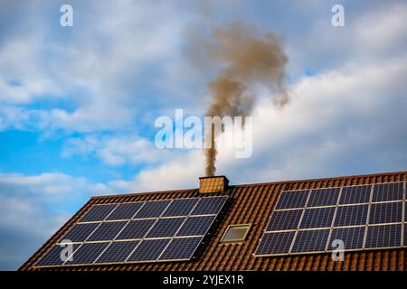 Fumo nero dal camino di una casa single-Family. Tetto coperto con pannelli solari. Tetto contro un cielo blu leggermente nuvoloso. Foto Stock