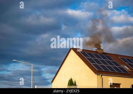 Fumo nero dal camino di una casa single-Family. Tetto coperto con pannelli solari. Tetto contro un cielo blu leggermente nuvoloso. Foto Stock