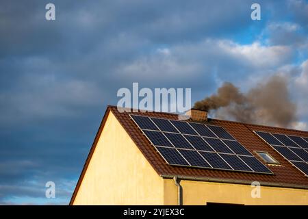 Fumo nero dal camino di una casa single-Family. Tetto coperto con pannelli solari. Tetto contro un cielo blu leggermente nuvoloso. Foto Stock