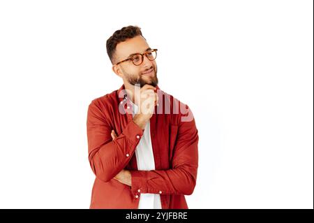 Attraente uomo arabo o caucasico con occhiali, vestito con una camicia rossa casual con barba, tenendo la mano su un mento, in piedi su sfondo bianco isolato, pensando a qualcosa e guardando lontano Foto Stock