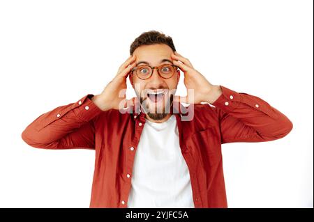 Eccitato giovane uomo caucasico o arabo con gli occhiali che tengono la testa in stupore, indossando una camicia casual rossa su uno sfondo bianco isolato. Espressivo, allegro e sorpreso Foto Stock