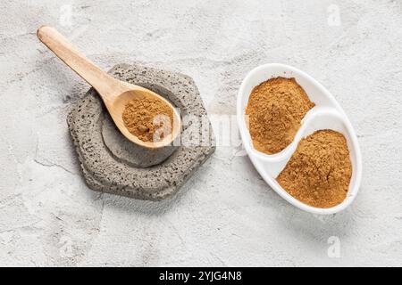 Un cucchiaio di legno pieno di cannella smerigliato si trova su una base di pietra grigia. Accanto ad esso, un piatto bianco diviso contiene due porzioni di spezia. Foto Stock