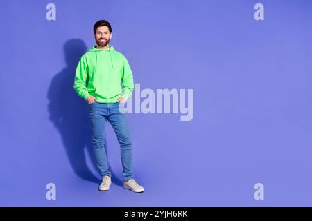 Foto a grandezza naturale di un giovane uomo che posa le tasche delle mani indossano abiti verdi alla moda isolati su sfondo viola Foto Stock