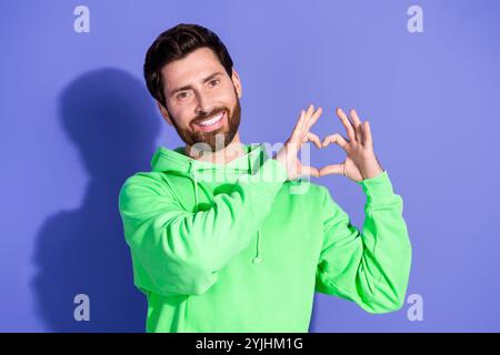 Foto ritratto di un bel fidanzato che sorride un uomo che indossa una felpa con cappuccio verde neon che mostra l'amore simbolo del cuore isolato su sfondo di colore viola Foto Stock