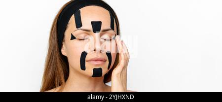 Ritratto di bellezza di una donna caucasica che applica nastri di kinesiologia nera sul viso. Foto Stock