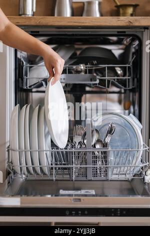 Vista frontale dalla mano femmina che rimuove la piastra pulita da una lavastoviglie caricata. Sfondo di una cucina moderna Foto Stock