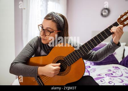 Donna che si concentra mentre suona la chitarra acustica nella sua camera da letto, godendo il suo hobby Foto Stock