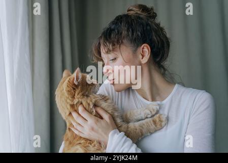 Giovane donna che coccola il suo adorabile gatto zenzero, esprime amore e affetto attraverso un tenero bacio, creando un momento di sentimento di connessione tra uomo e animale Foto Stock