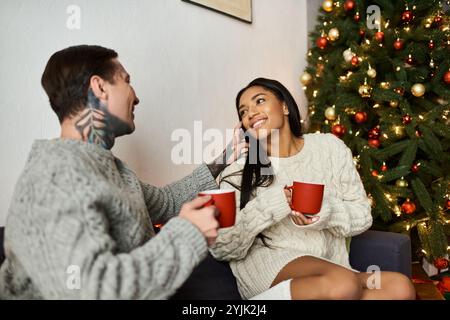 Una coppia si diverte a stare insieme, ridendo e sorseggiando bevande calde vicino a un albero di Natale festivo. Foto Stock