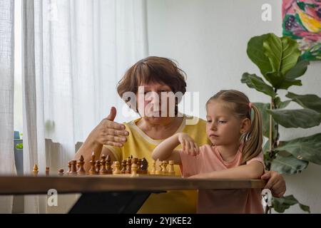 Una ragazza allegra gioca a scacchi con la nonna a tavola, creando preziosi ricordi di famiglia. Foto di alta qualità Foto Stock