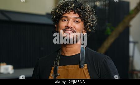 Felice sorridente gioioso giovane indiano arabo uomo lavoratore di caffè in grembiule maschio ragazzo cameriere aspetto più pulito pensare a distanza girando guardando alla fotocamera amichevole Foto Stock