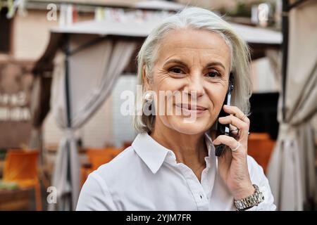 Una donna graziosa con i capelli grigi sorride mentre parla al telefono in un ambiente incantevole all'aperto. Foto Stock