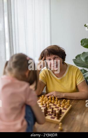 Una ragazza allegra gioca a scacchi con la nonna a tavola, creando preziosi ricordi di famiglia. Foto di alta qualità Foto Stock