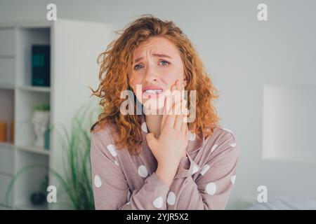 Giovane donna dai capelli rossi in pigiama a pois che esprime preoccupazione e disagio in un'accogliente camera da letto con luce naturale Foto Stock