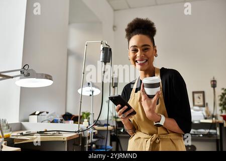 Una donna allegra tiene in mano una tazza di caffè e un telefono mentre è circondata da strumenti per la creazione di gioielli. Foto Stock