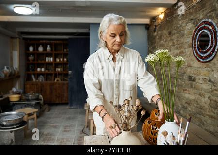 Una bella donna matura organizza con cura delicati fiori in vasi su un rustico tavolo in legno. Foto Stock