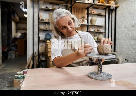 Una donna matura di talento si concentra intensamente sulla creazione di un pezzo di ceramica unico nel suo laboratorio. Foto Stock