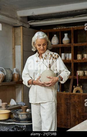 La donna matura ha in mano un vaso unico pieno di fiori secchi, sorridendo dolcemente nel suo studio. Foto Stock