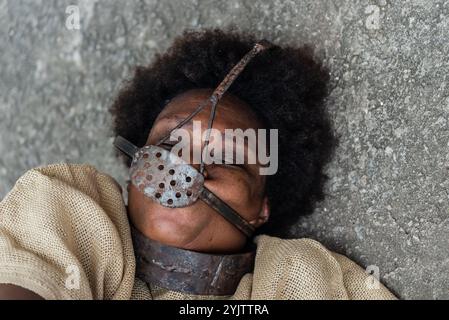 Ritratto ravvicinato del volto di una donna nera con una maschera di ferro sopra la bocca e il collo distesi sul pavimento con un'espressione di dolore. Schiavitù a Bra Foto Stock