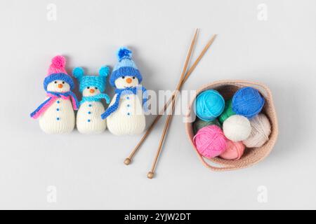 Tre giocattoli di pupazzo di neve a maglia in cappelli e sciarpe blu e rosa, aghi per maglieria e cesto con palline colorate di filo su sfondo grigio. Regalo per le vacanze invernali. Vista dall'alto, disposizione piatta. Foto Stock