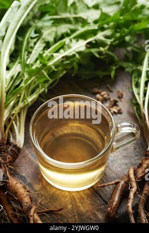 Tè alle erbe in una tazza di vetro con radici di dente di leone fresche e foglie su un tavolo di legno Foto Stock