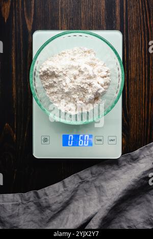 Pesatura della farina multiuso su una bilancia digitale da cucina: Vista dall'alto della farina di frumento sbiancata in un recipiente di vetro pesato su una bilancia alimentare Foto Stock