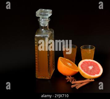 Liquore arancia-pompelmo con spezie su sfondo nero, alcool fatto in casa in una ciotola di cristallo accanto a frutta matura e cannella con anice. Primo piano. Foto Stock