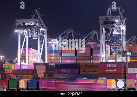 Wilmington, California, USA - 16 luglio 2021: Gru scaricano container intermodali da una nave nel porto di Los Angeles, mentre i semi-camion trasportano t Foto Stock