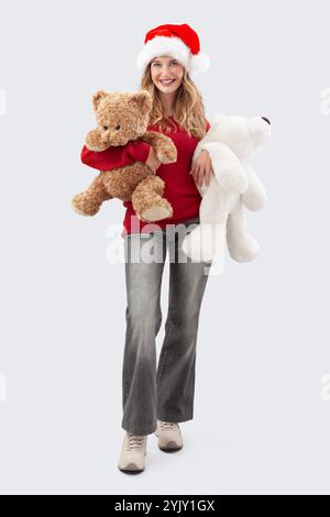 Buon Natale, la giovane donna felice e gioiosa che indossa un cappello di Babbo Natale sorride mentre tiene con grande tenerezza gli orsacchiotti, abbracciandoli saldamente Foto Stock