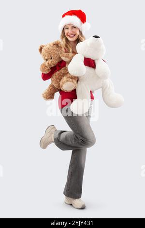 Buon Natale, la giovane donna felice e gioiosa che indossa un cappello di Babbo Natale sorride mentre tiene con grande tenerezza gli orsacchiotti, abbracciandoli saldamente Foto Stock