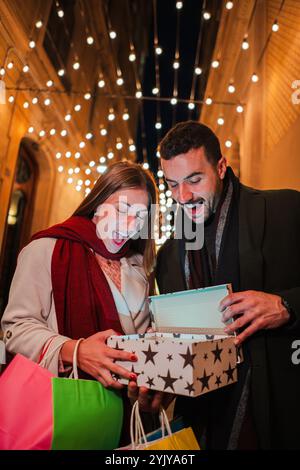 Verticale. Giovani coppie che sfoggiano un regalo di Natale all'aperto di notte, sorridono di emozioni sotto le luci delle feste in una strada decorata per le feste, godendo lo spirito di dare durante le feste. Foto di alta qualità Foto Stock