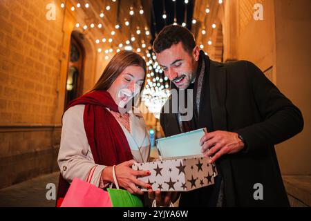 Bella giovane coppia che apre un regalo di Natale all'aperto di notte, sorridendo di emozioni sotto le luci festive in una strada decorata per le feste, godendo lo spirito di dare durante le feste. Foto di alta qualità Foto Stock