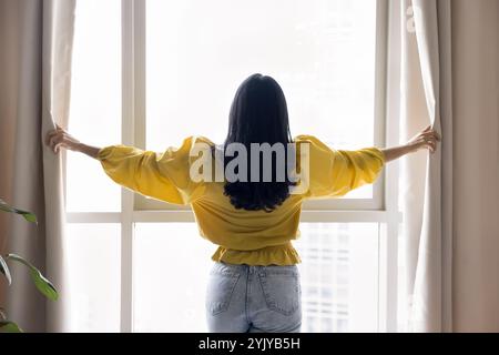 Retro di una giovane donna in abiti casual che aprono tende Foto Stock