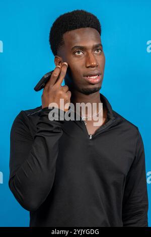 Giovane afroamericano che utilizza auricolari wireless con tecnologia a conduzione ossea per ascoltare musica o podcast su sfondo blu Foto Stock