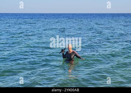 Nuotatrice senior con capelli corti e bagnati grigi in costume da bagno nero chiuso tiene la maschera e il boccaglio mentre si trova in acqua blu, esprimendo gioia al sole Foto Stock