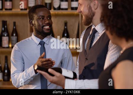 Sommelier afroamericano sorridente ampiamente mentre comunica con i suoi colleghi birazziali prima di degustare il vino Foto Stock