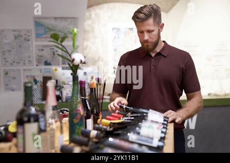 Primo piano medio del lavoratore di una cantina caucasica che regola una bottiglia di vino rosso, si trova vicino a una cabina di legno con vini rossi e bianchi Foto Stock