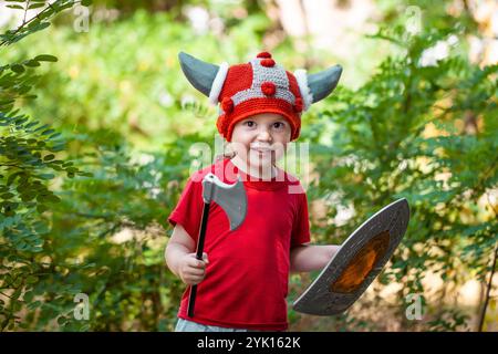 Ragazzo carino che gioca con la spada e lo scudo fuori. Idee per giochi di ruolo per bambini. Cappello vichingo fai da te. Stile di vita Foto Stock