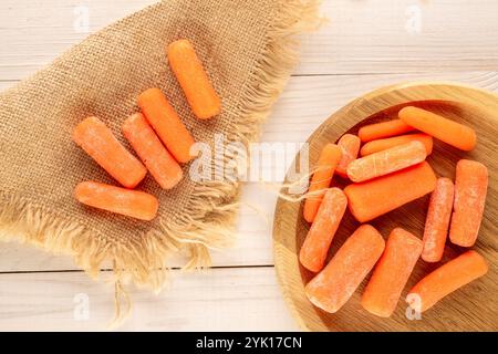 Piccole carote pelate con utensili in legno e tovagliolo in iuta su un tavolo in legno, vista dall'alto. Foto Stock