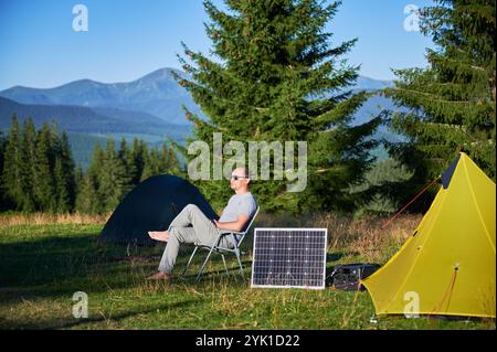 Uomo che utilizza un pannello solare fotovoltaico per caricare una centrale elettrica portatile all'aperto in campeggio ecologico. Il turista maschile siede rilassato su una sedia vicino alle tende. Sullo sfondo alti pini e cielo azzurro. Foto Stock