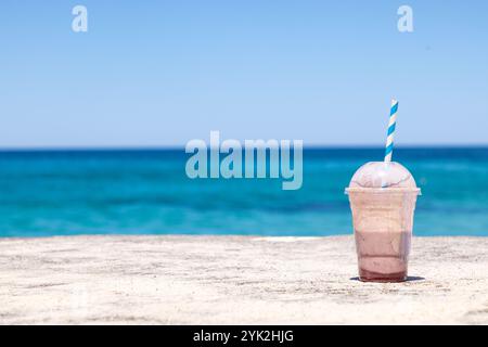 Frullato di cioccolato mezzo pieno con paglia blu e bianca poggia su una superficie sabbiosa vicino al mare a Bronte Beach, Sydney, Australia. Foto Stock