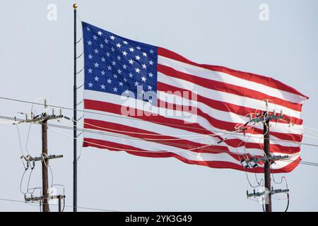 Bandiera DEGLI STATI UNITI che sventola al vento (a distanza di sicurezza) dietro i pali di energia elettrica, nella contea di Bexar, Texas, il 5 giugno 2024. Fornire elettricità affidabile e a prezzi accessibili è essenziale per sostenere il benessere economico e la qualità della vita di tutti i residenti rurali della nazione. U/S. Il programma elettrico del Department of Agriculture (USDA) Rural Development (RD) Rural Utilities Service (RUS) fornisce leadership e capitale per mantenere, espandere, aggiornare e modernizzare le vaste infrastrutture elettriche rurali americane. Foto Stock