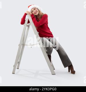 Buon Natale, felice donna sorridente seduta su una scala di legno vintage prima di decorare l'albero di Natale guardando la macchina fotografica con il cappello di Babbo Natale, io Foto Stock