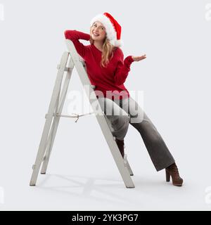 Buon Natale buon Natale, felice donna sorridente seduta su una scala di legno vintage che mostra la mano prima di decorare l'albero di Natale guardando nel Foto Stock