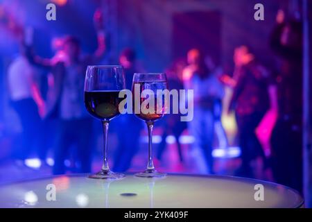 Due bicchieri sul tavolo al nightclub con sfondo vivace. L'energica scena di festa presenta folla danzante e luci colorate. Foto Stock