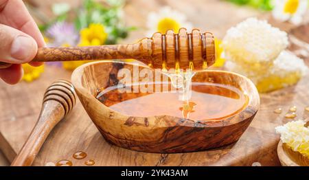 Miele che gocciola dal cucchiaio di legno in una ciotola di legno con miele, i fiori primaverili sono su un tavolo di legno vicino. Foto Stock